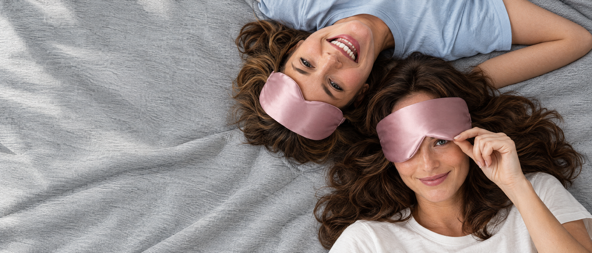 Two friends lying head to head on a grey blanket wearing pink silk sleep masks